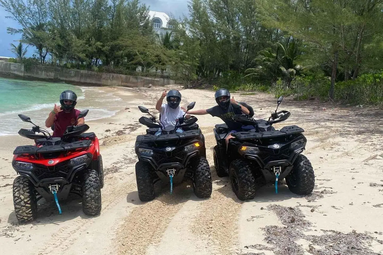 Three riders in helmets on red and black ATVs parked on a sandy track beside the sea