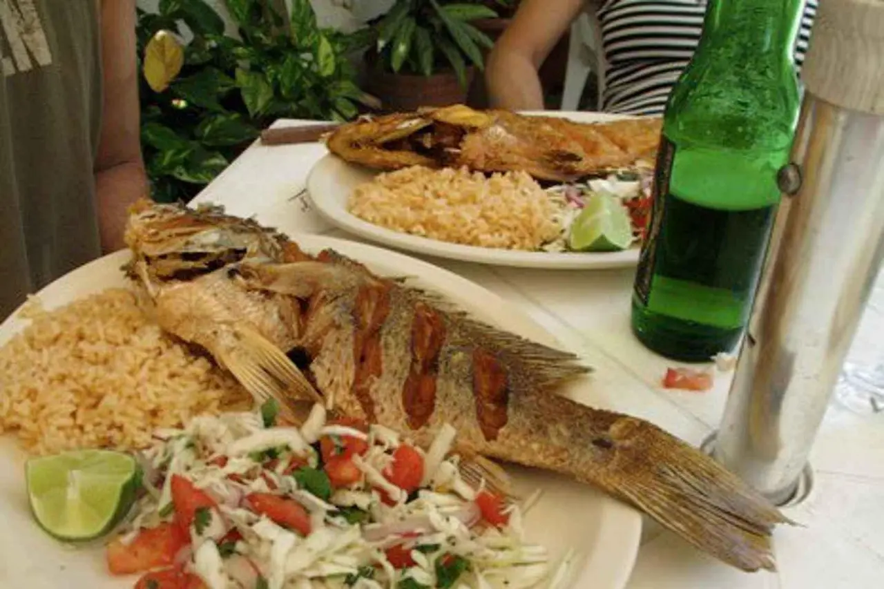 Two plates of whole fried fish with rice, cabbage salad and a lime wedge beside a green bottle on a white table