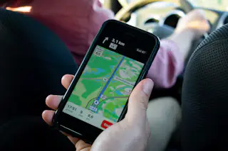 A hand holding a smartphone that shows a turn-by-turn navigation map, with a driver's arm on the wheel behind it