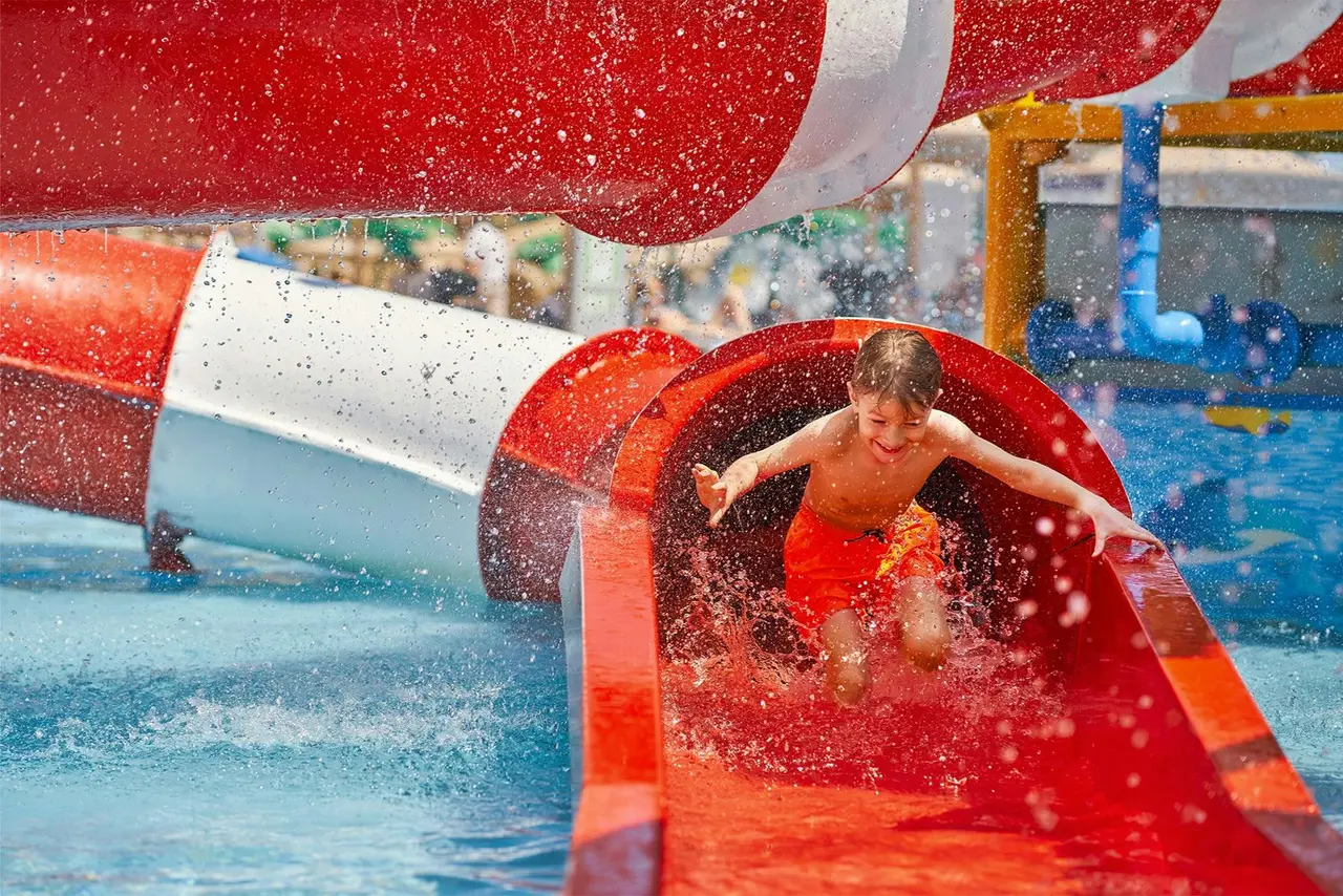 A boy in orange swim trunks shooting down a red-and-white waterslide as water sprays around him